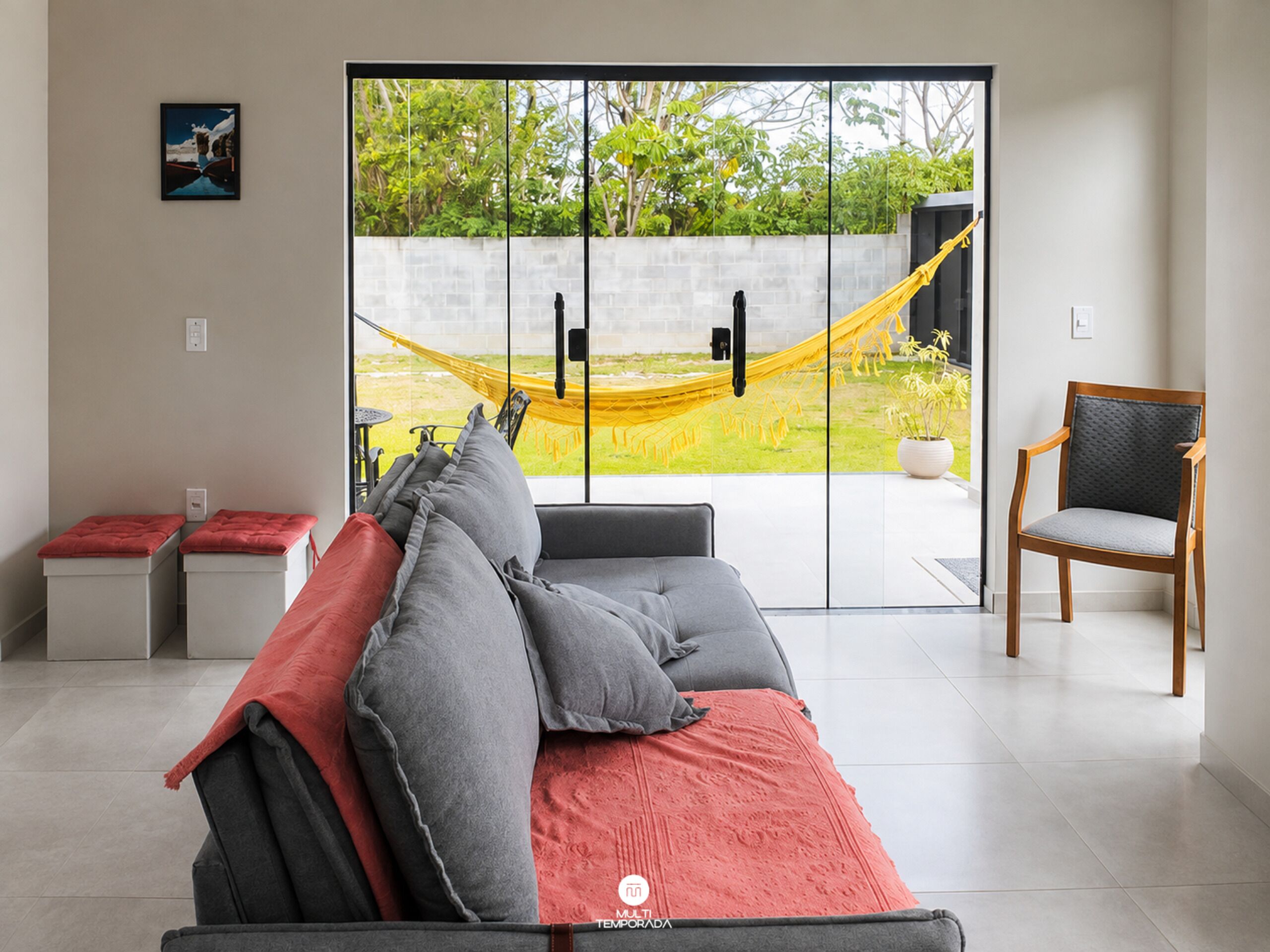 A modern living room with a sofa, a chair, a hammock, and a view of a garden through a glass door.