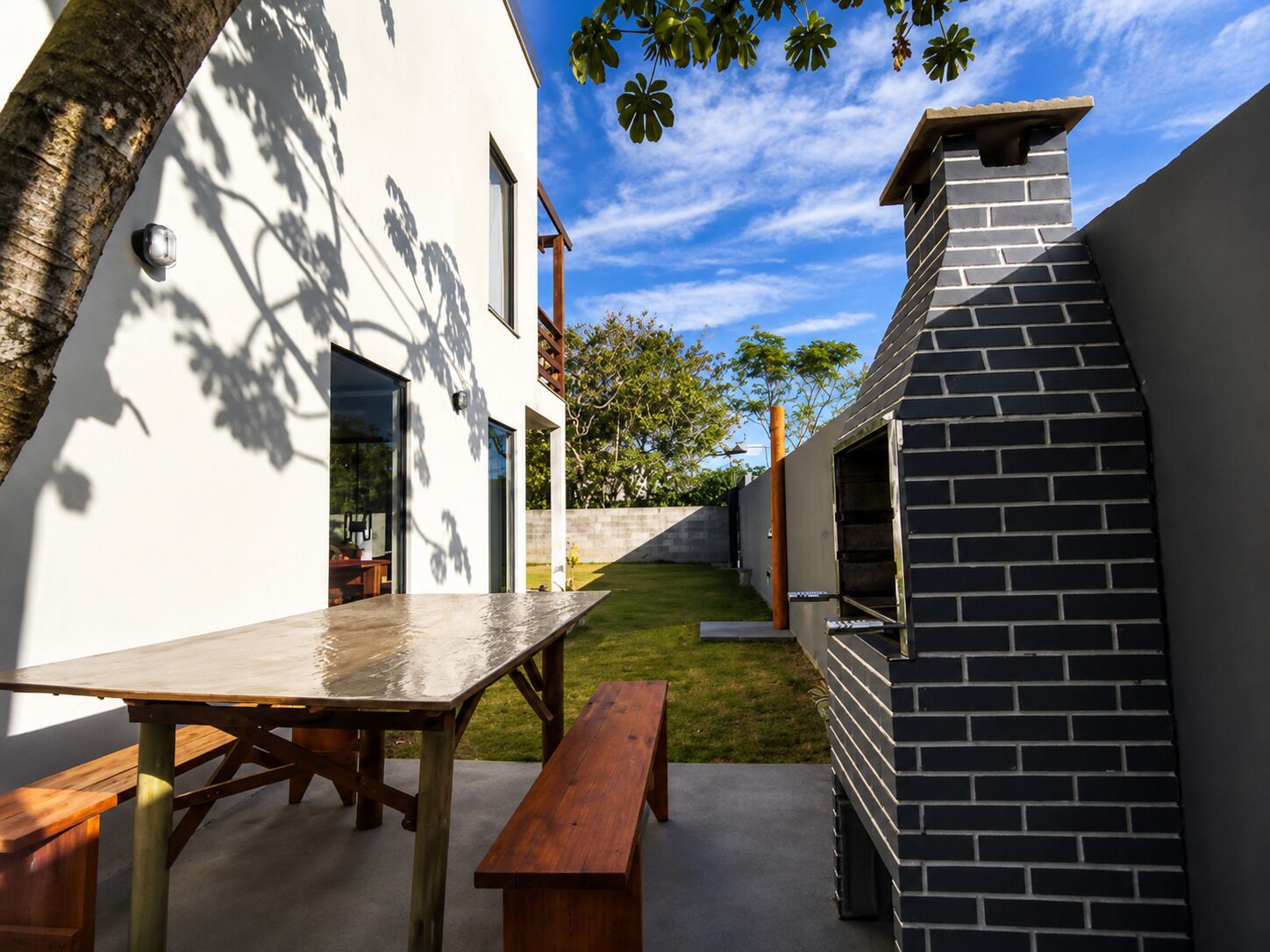 A modern outdoor patio with a wooden table and benches, a brick barbecue grill, and a white building with large windows.