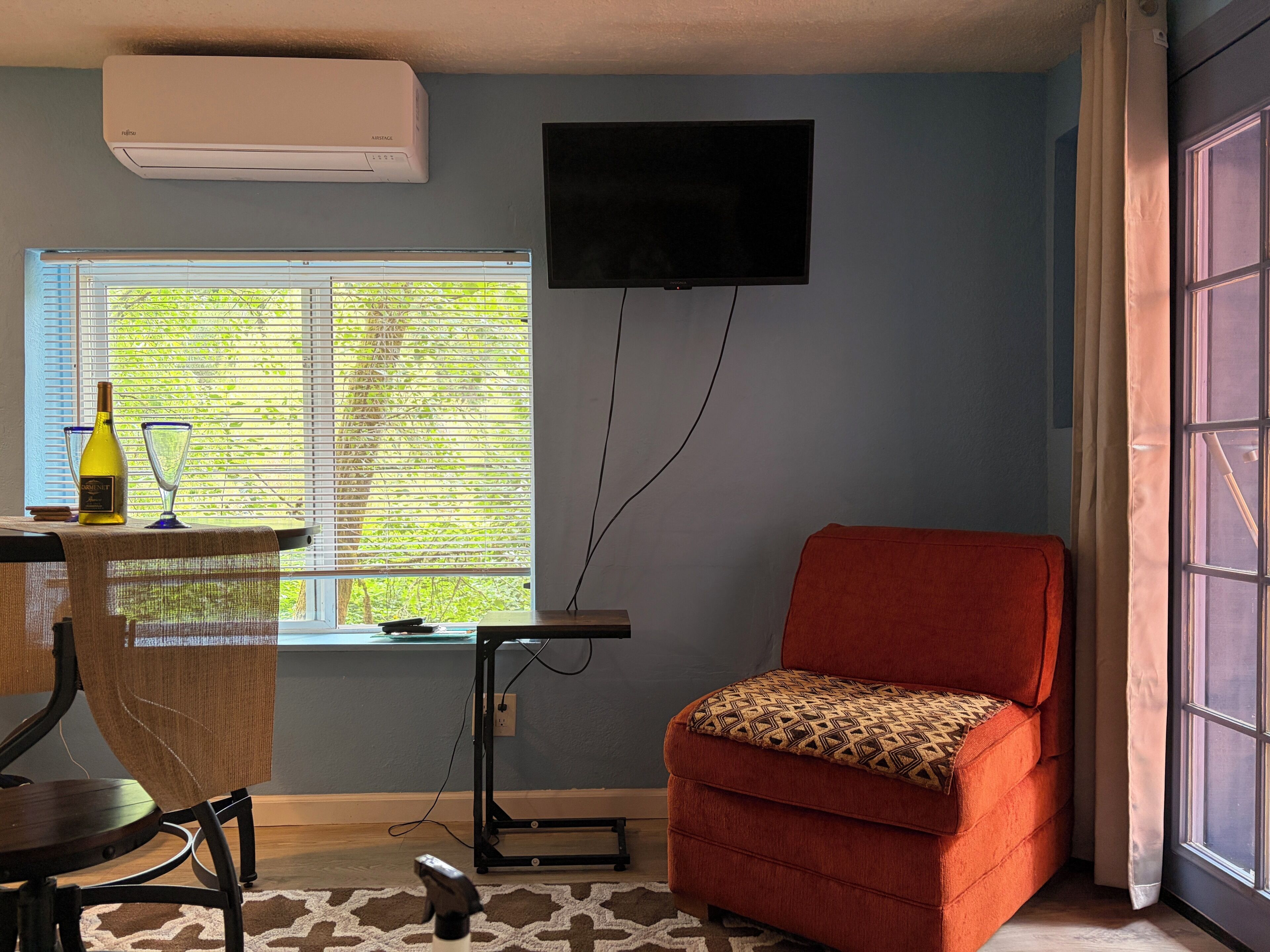 A room with a TV mounted on the wall, a window with blinds, a red chair, a table with a bottle and glasses, and an air conditioning unit.