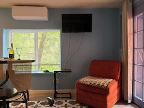 A room with a TV mounted on the wall, a window with blinds, a red chair, a table with a bottle and glasses, and an air conditioning unit. - Cozy studio apartment in beautiful Guerneville with AC overlooking the creek (Guerneville)