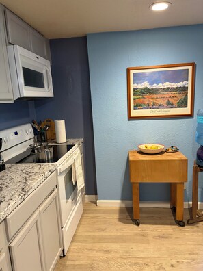 A kitchen with white cabinets, a microwave, a stove, and a wooden table with a bowl of fruit. - Cozy studio apartment in beautiful Guerneville with AC overlooking the creek (Guerneville)