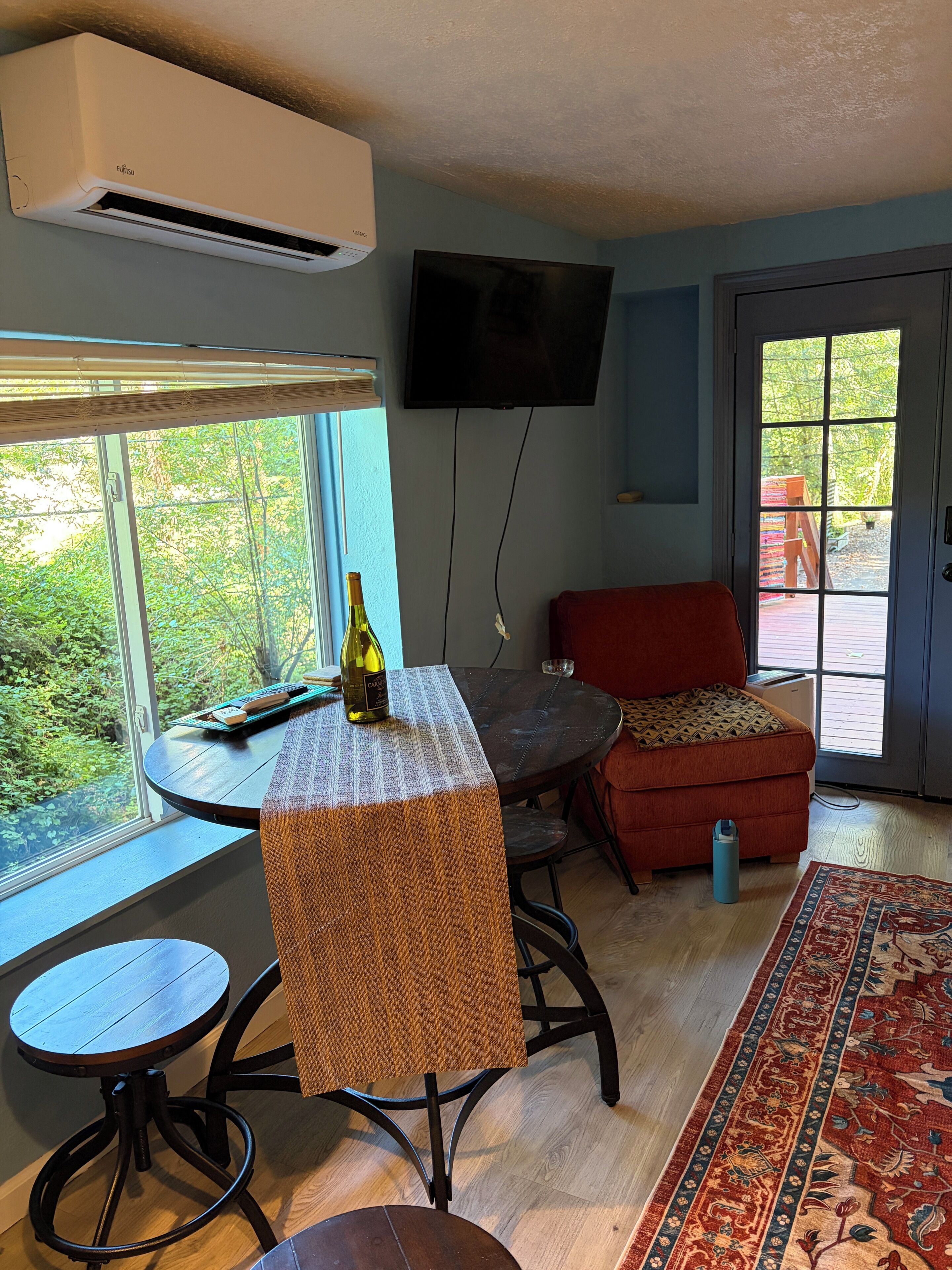 A room with a dining table, a red sofa, a TV mounted on the wall, and a window with a view of greenery.