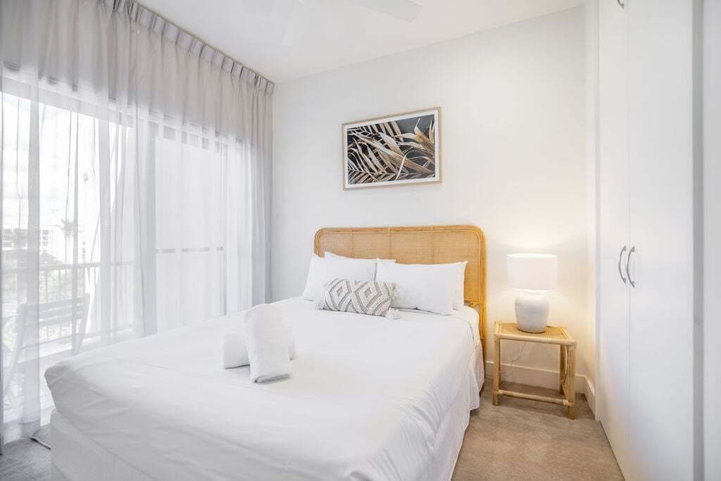Una camera d'albergo con un letto grande, un comodino con una lampada, un quadro incorniciato al muro e tende leggere alla finestra.