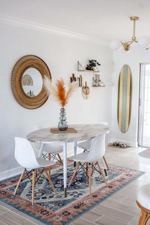 A dining area with a round table, white chairs, a patterned rug, and a surfboard decoration on the wall. - Bonita Shores Beach House Close to Doc's, Barefoot Beach, Naples +Dog-Friendly! (Bonita Springs)