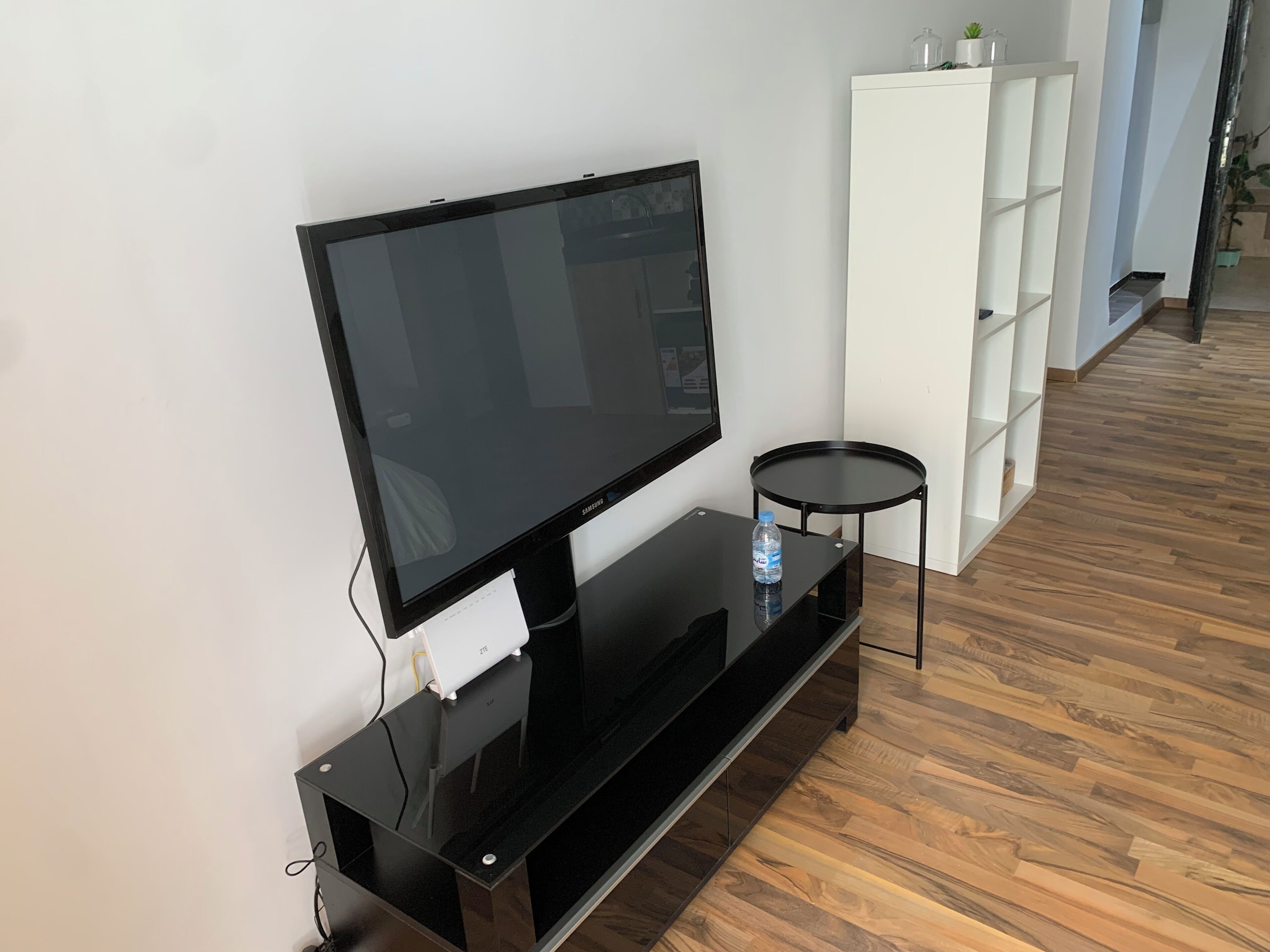 A flat-screen TV on a black stand, a small round table, and a white shelving unit in a room with wooden flooring.