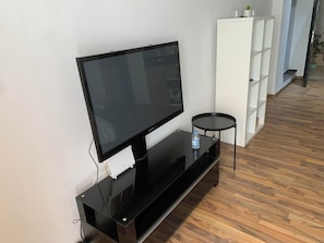 Ein Flachbildfernseher auf einem schwarzen Ständer, ein kleiner runder Tisch und eine weiße Regaleinheit in einem Zimmer mit Holzboden.