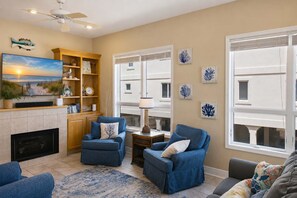 A living room with a fireplace, two blue armchairs, a coffee table, a TV, and a bookshelf. - Sea Forever SGI Beach Front 5 Bedroom  Hot tub Pool (Saint George Island)