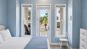 Een slaapkamer met een bed, een stoel, een dressoir en een balkon met uitzicht op palmbomen en gebouwen.