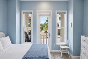 Ein Schlafzimmer mit Bett, einem Sessel, einem Schminktisch und einem Balkon mit Blick auf Palmen und Gebäude.