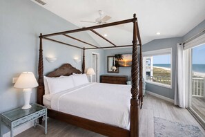Un lit à baldaquin avec une structure en bois et une literie blanche, une table d’appoint en verre avec une lampe, un ventilateur de plafond et une vue sur la plage par une grande fenêtre.
