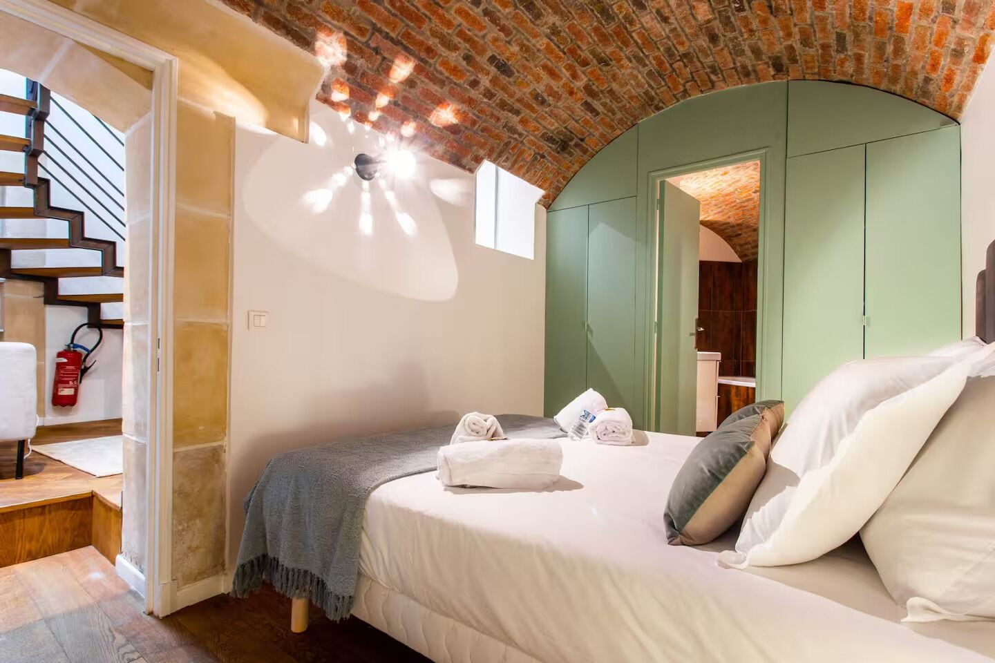 Une chambre avec un plafond en briques, un lit recouvert de draps blancs, une porte d’entrée en arc de cercle de couleur verte et un escalier équipé d’un extincteur.