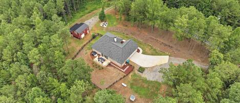 A house surrounded by dense forest, with a large driveway and a covered outdoor area.