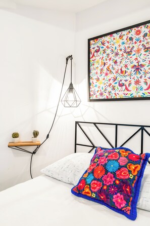A bedroom with a bed, a hanging light fixture, a wall-mounted shelf with two potted plants, and a colorful floral painting above the bed. - Loft in the Historic Center of the City (Ciudad de México)