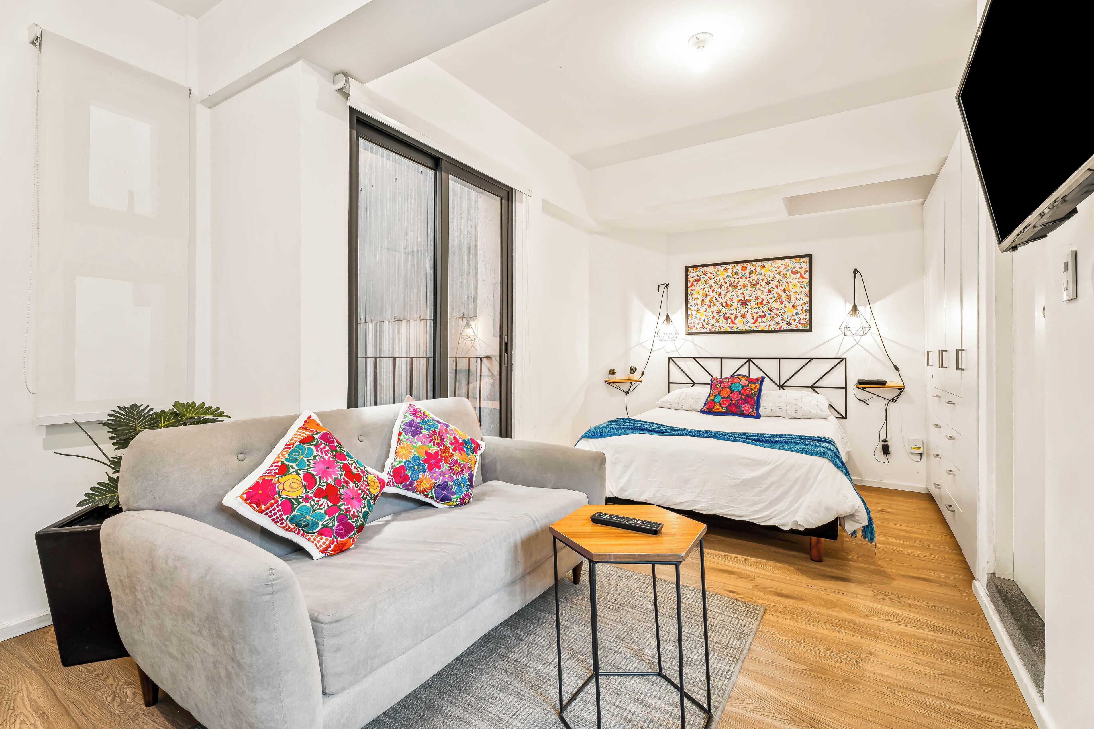 Una camera d'albergo moderna con un divano grigio, un tavolino, un letto con coperta blu e un'opera d'arte colorata appesa al muro.