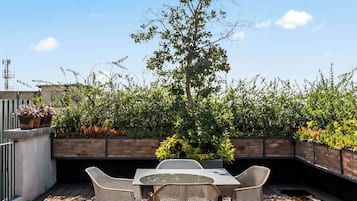 An outdoor patio with a table and chairs, surrounded by greenery and a clear sky.