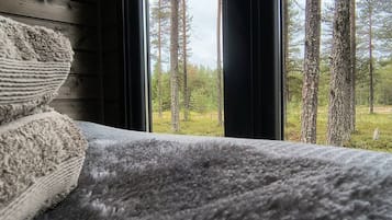 Een bed met een grijze deken en uitzicht op een bos door een raam.