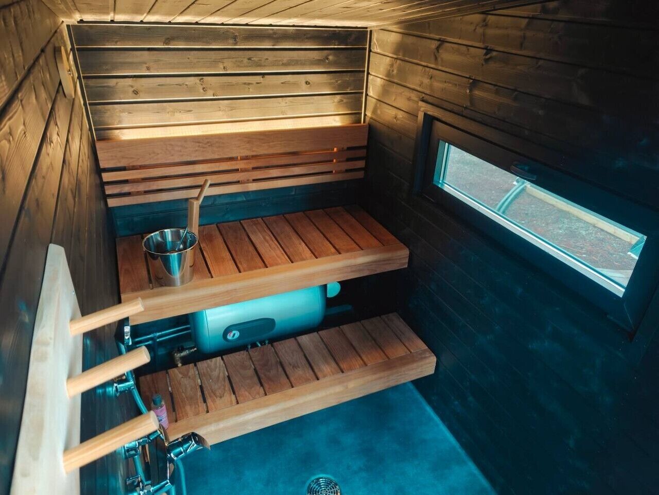 A sauna with wooden benches, a metal bucket, and a window.
