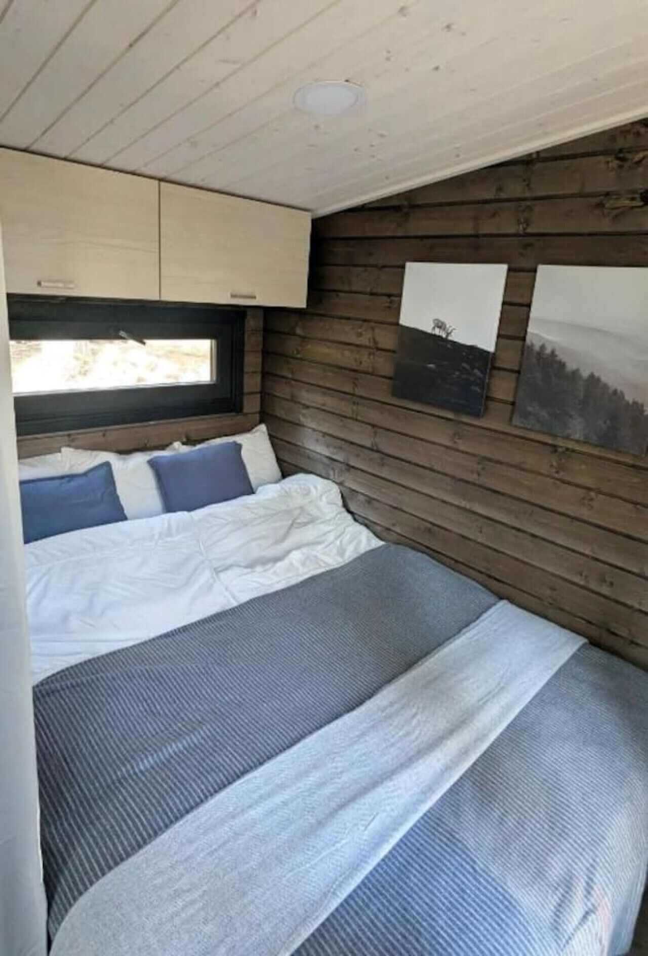 A bedroom with a bed, pillows, a window, wooden walls, and artwork.
