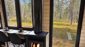 A cozy room with a wood-burning stove, a dining table with chairs, and large windows offering a view of a forest.