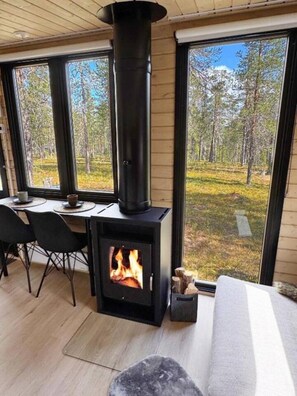 Una habitació acollidora amb una llar de llenya, una taula de dinar amb cadires i grans finestres que donen vistes a un bosc.