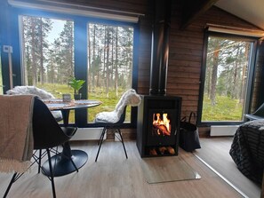 A cozy room with a wood-burning stove, a dining table with chairs, and large windows overlooking a forest.