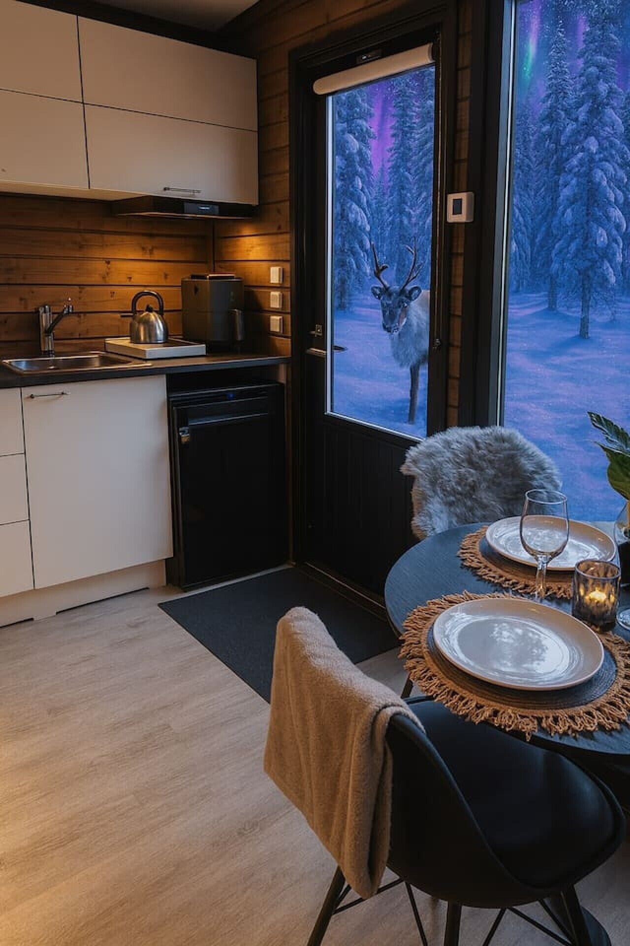 Eine moderne Küche mit Einbauherd, Wasserkocher und Kühlschrank. Es gibt einen Essbereich mit einem für zwei Personen gedecktem Tisch, einem Stuhl und Blick auf einen verschneiten Wald durch ein großes Fenster.