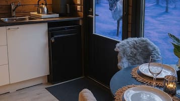 Eine moderne Küche mit Einbauherd, Wasserkocher und Kühlschrank. Es gibt einen Essbereich mit einem für zwei Personen gedecktem Tisch, einem Stuhl und Blick auf einen verschneiten Wald durch ein großes Fenster.
