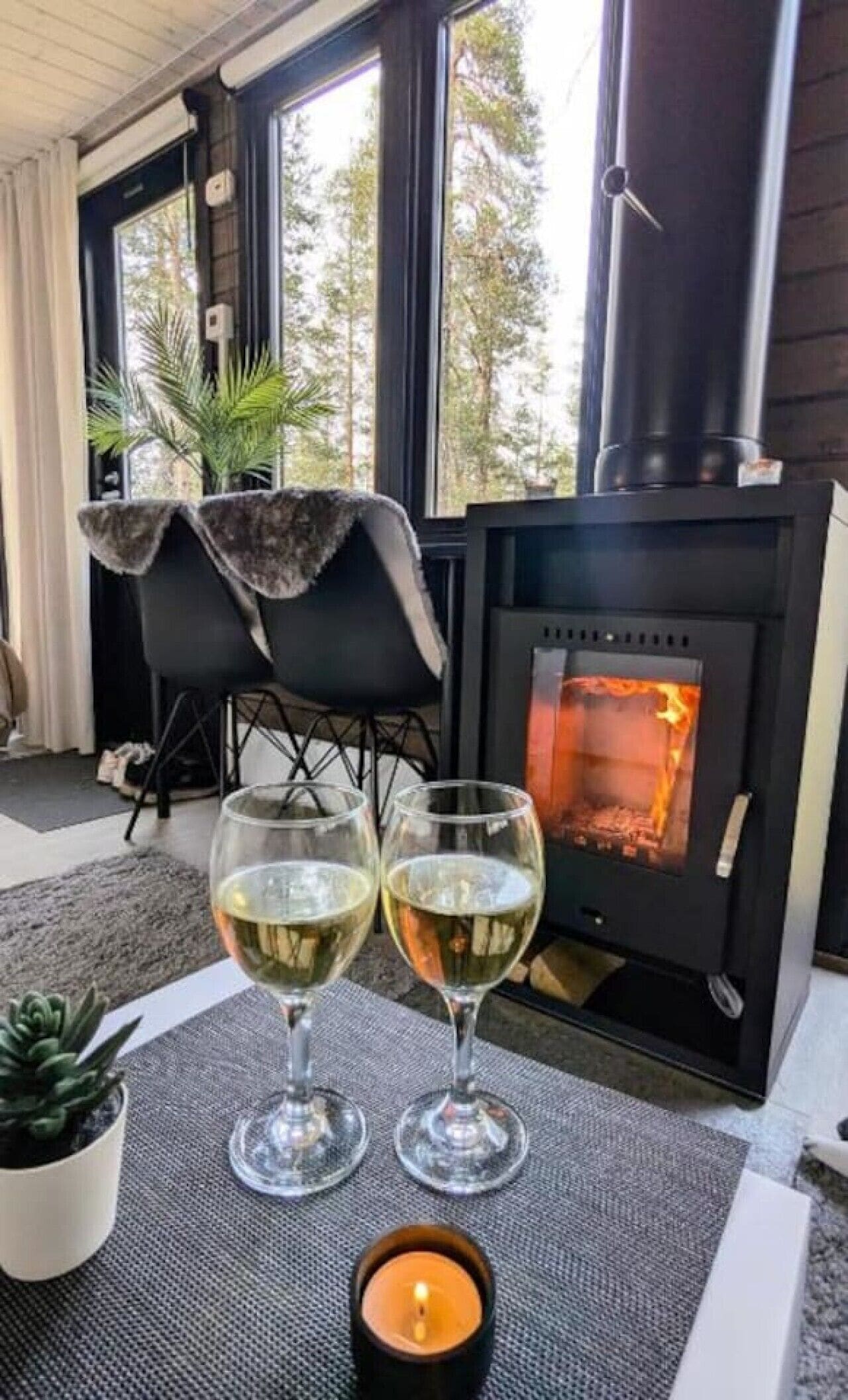 Um quarto aconchegante com lareira, dois copos de vinho branco, uma vela e uma cadeira.