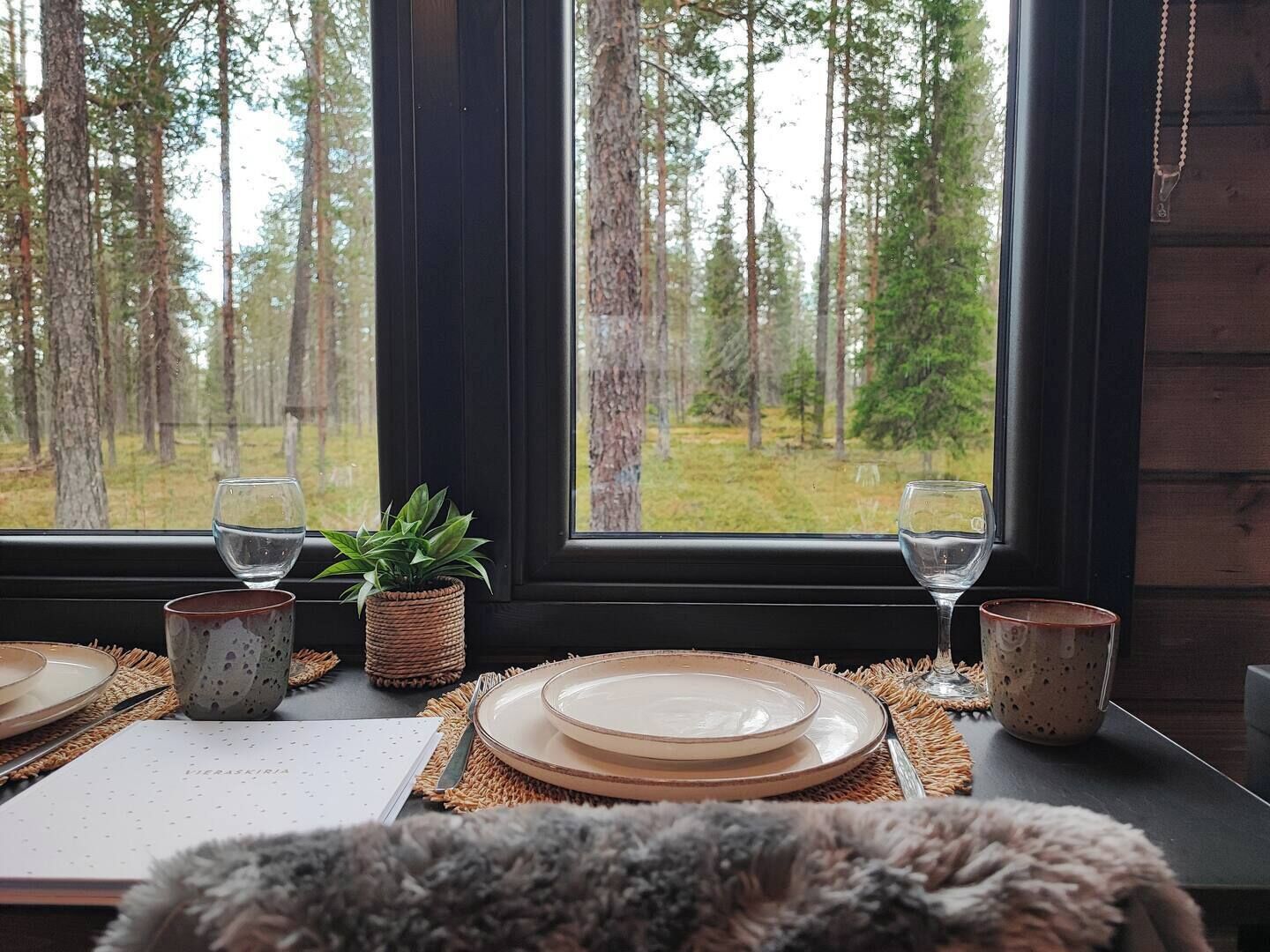 A dining table set with plates, glasses, and a menu, with a view of a forest through a window.