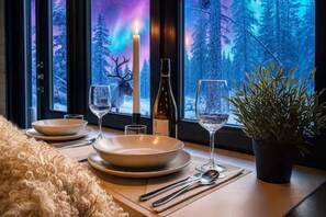 Mesa lista con platos, vasos, una vela y una botella, con vista a un bosque desde la ventana.