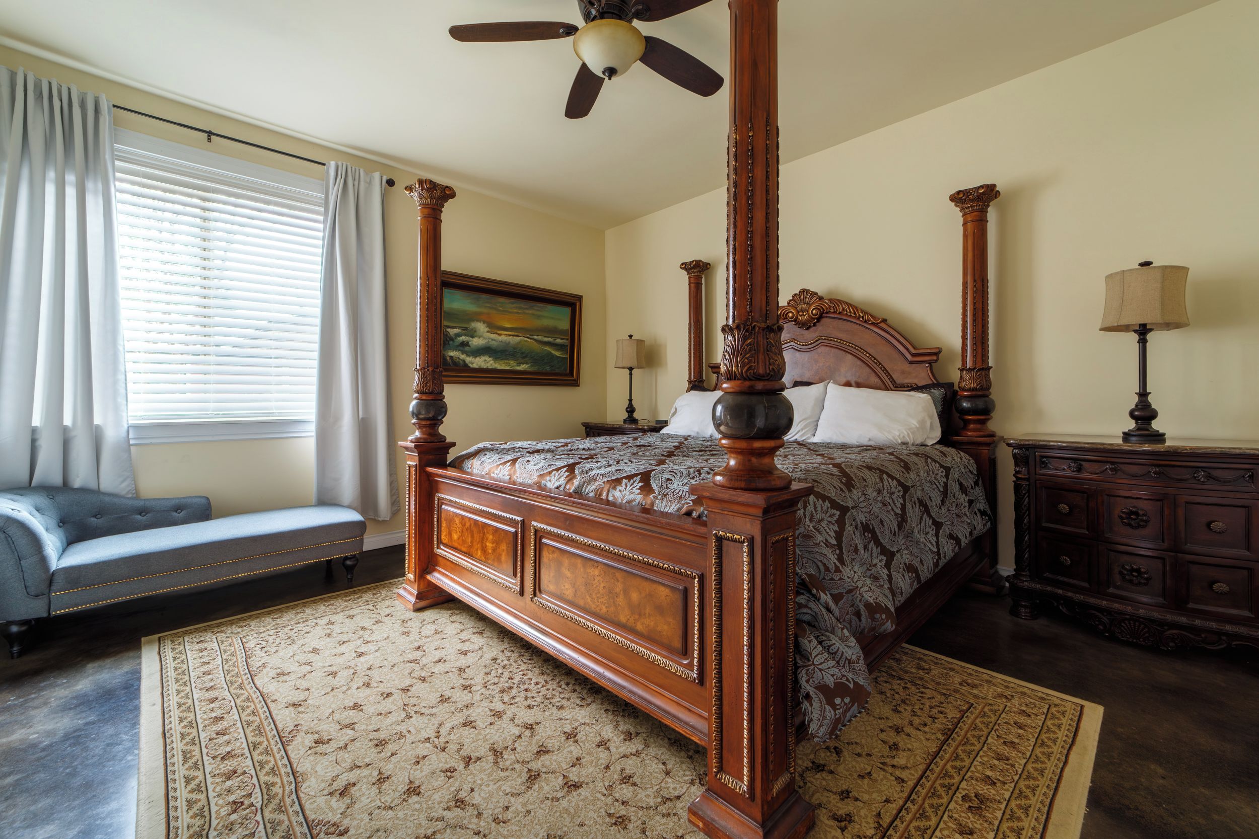 A four-poster bed with a headboard, a bench, a painting, and a lamp on a dresser.