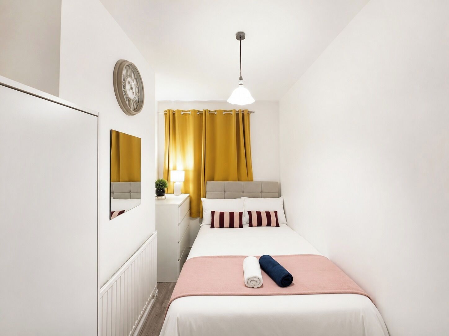 Un dormitori amb un llit, una taula de nit, una làmpada, un mirall i una finestra amb cortines.