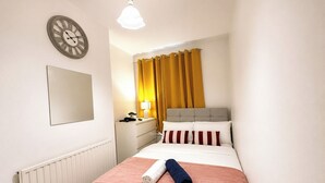 Ein Hotelzimmer mit einem Bett, einem Nachttisch, einer Lampe, einer Uhr und einem Fenster mit Vorhängen.