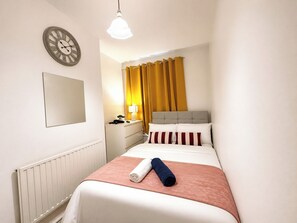 Ein Hotelzimmer mit einem Bett, einem Nachttisch, einer Lampe, einer Uhr und einem Fenster mit Vorhängen.