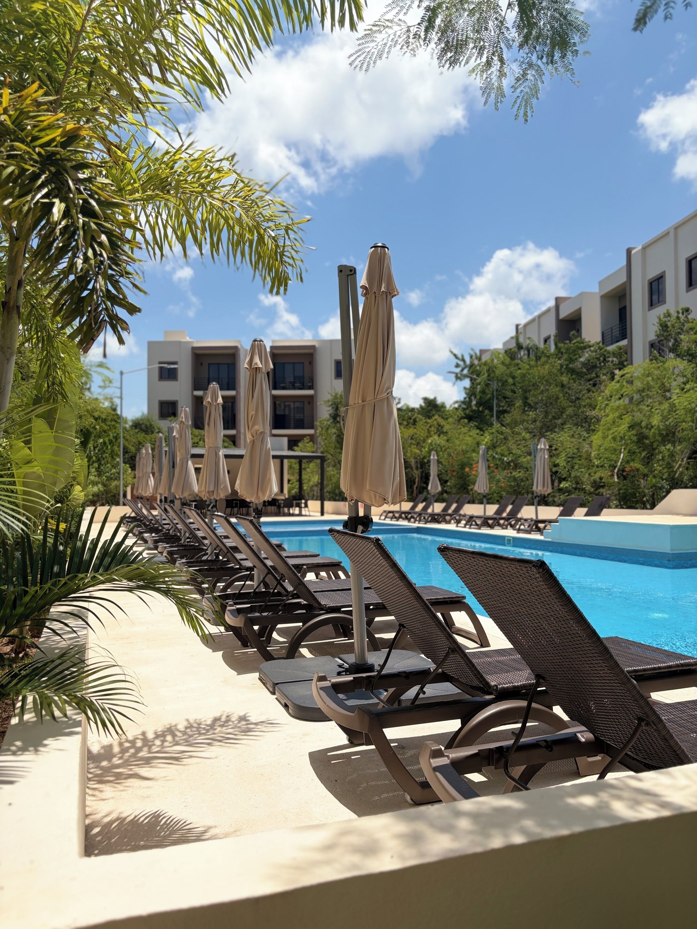A poolside area with lounge chairs and umbrellas, a clear blue pool, and a building with balconies in the background.