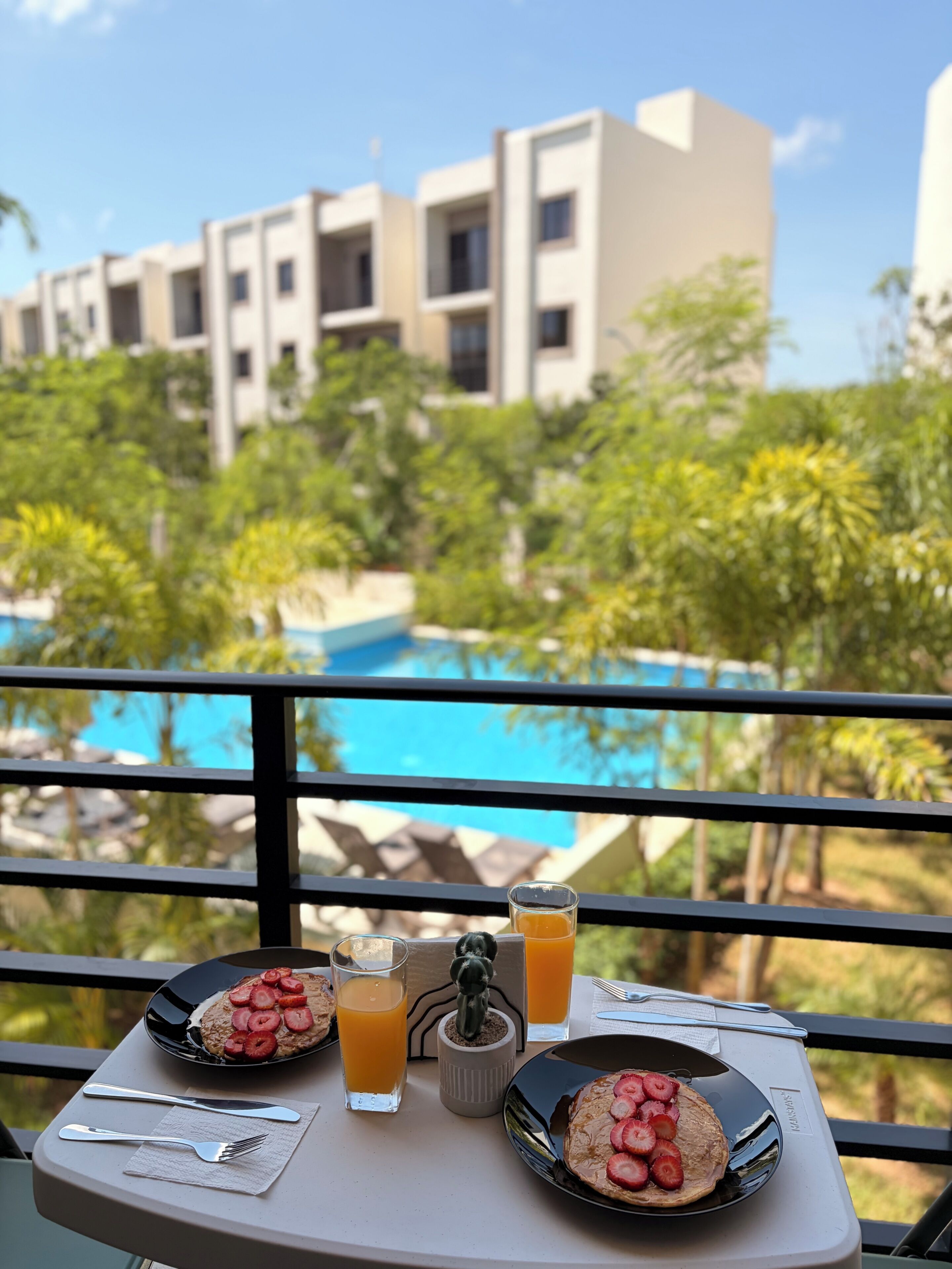 A dining table with breakfast items and a view of a swimming pool and buildings in the background.