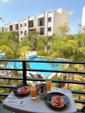 A dining table with breakfast items and a view of a swimming pool and buildings in the background.