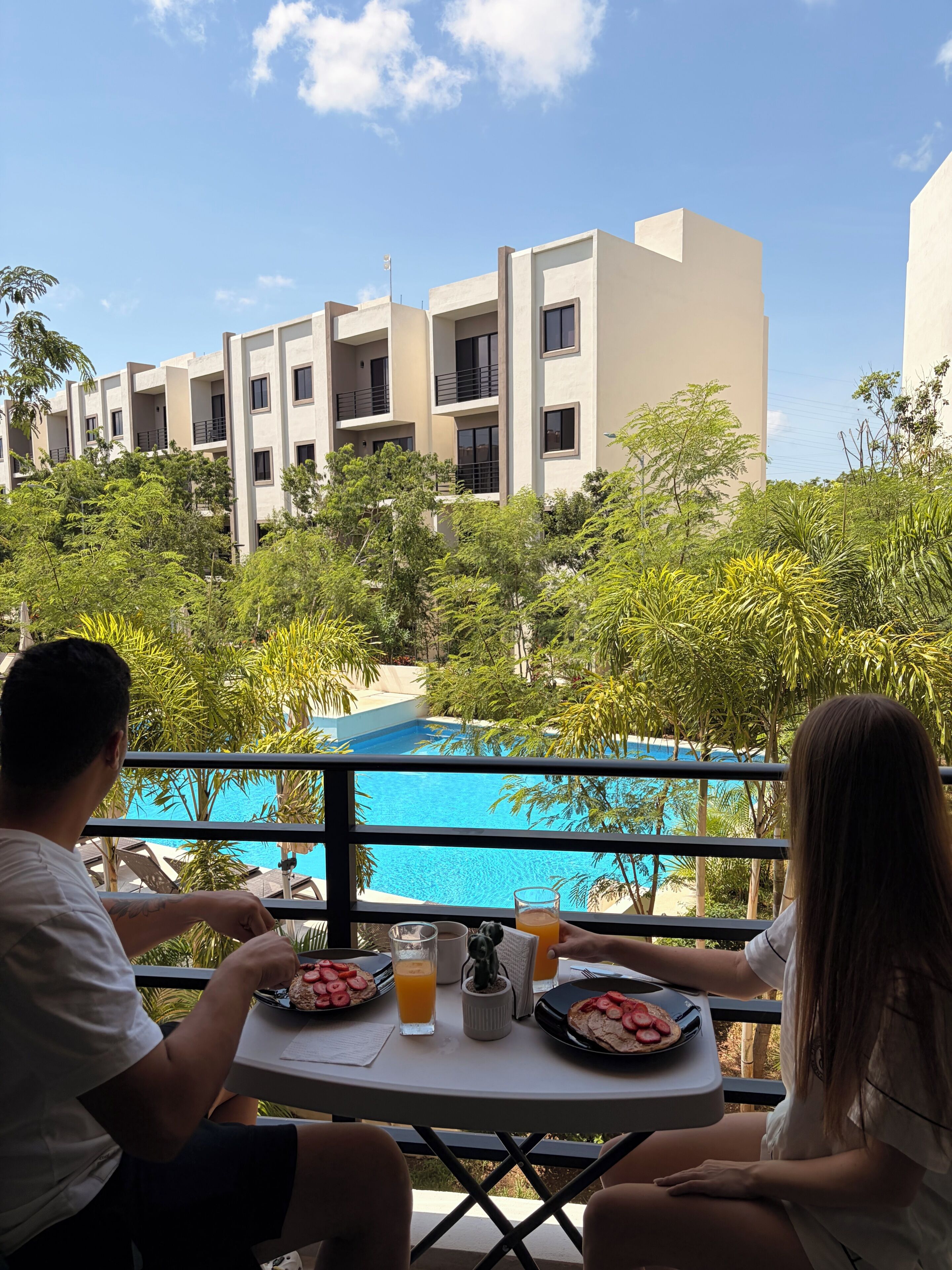 A dining table with breakfast items and a view of a swimming pool and buildings.