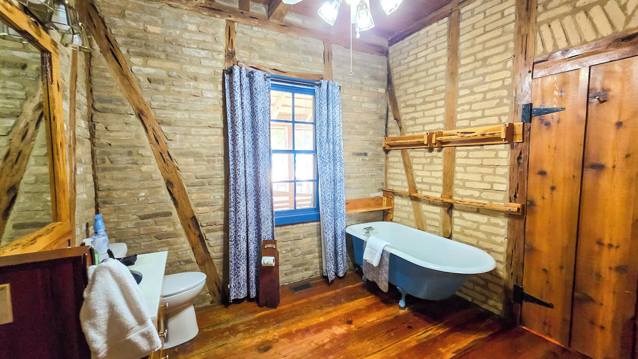Een landelijke badkamer met een houten vloer, een wit toilet, een blauw bad en een houten deur.
