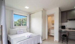 Hotellihuone, jossa on sänky, ikkuna verhoilla, näkymä puistoon, pieni keittiö ja kylpyhuone.
