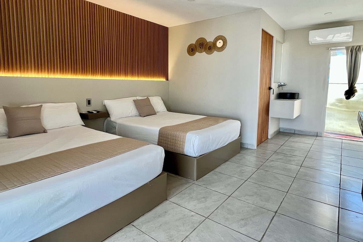 A hotel room with two beds, a wooden headboard, a wall-mounted air conditioning unit, and a bathroom with a sink and mirror.