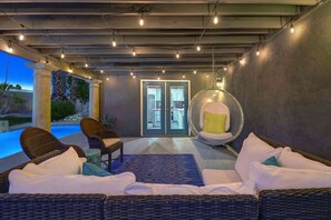 A covered outdoor seating area with wicker furniture, a hanging chair, and a view of a pool and landscape. - Navigate Stays Pool, Spa & Pet-Friendly PS Home (Palm Springs)