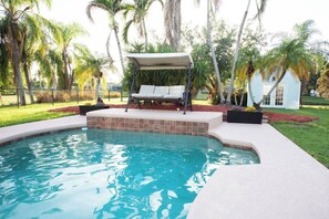 A swimming pool with a sun lounger and a covered seating area, surrounded by palm trees and a house in the background.