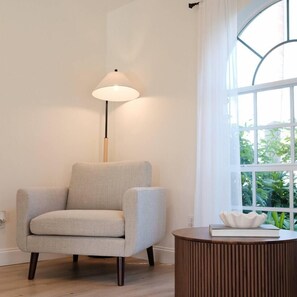 A room with a light-colored armchair, a round wooden side table, a floor lamp, and a large window with a view of greenery.