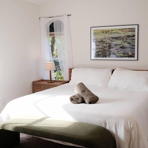 A neatly made bed with white linens and a rolled towel, a bedside table with a lamp, and a framed landscape painting on the wall.