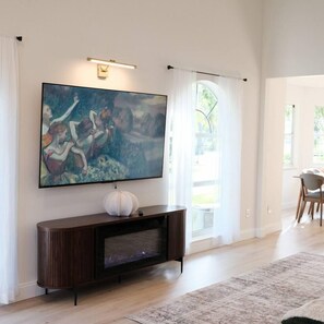 Een woonkamer met een grote flatscreen-televisie aan de muur, een donkere houten dressoir eronder en een groot raam met dunne gordijnen die natuurlijk licht doorlaten.