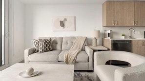 Ein modernes Wohnzimmer mit Sofa, Sessel, Couchtisch und einem Essbereich mit Lampe und Kaffeemaschine.