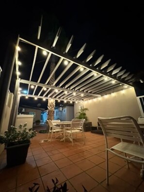 Ein Außenbereich mit Terrasse, einer Pergola, Fliesenböden und Gartenmöbeln.