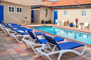 A poolside area with lounge chairs, a pool, and a building with blue doors and windows. - Escape w/ Pool · King Bed · Near Palm Beach #7 (Aruba)