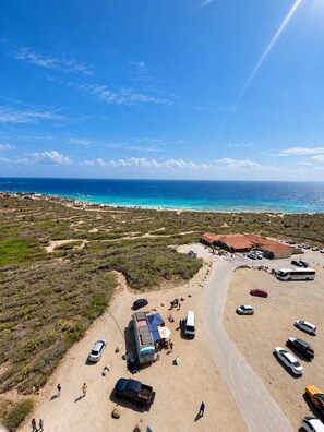 An aerial view of a coastal area with a parking lot, beach, and buildings. - Aruba Studio w/ Pool · King Size · Near Beach #13 (Noord)